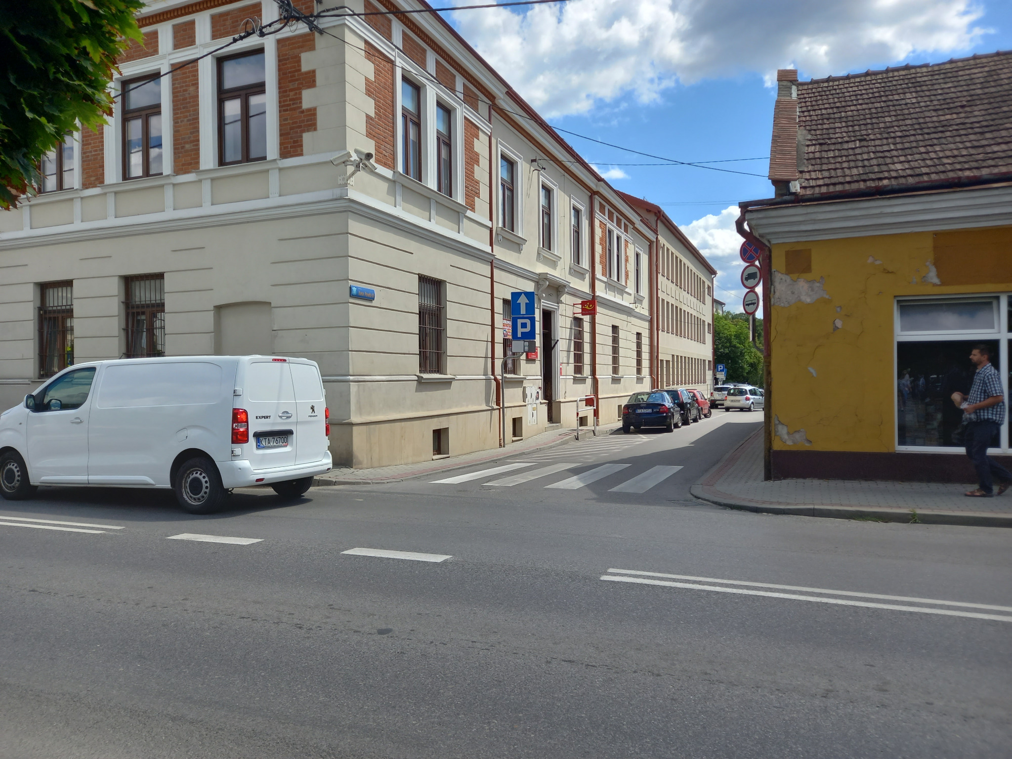 Biały samochód dostawczy marki Peugeot Expert zaparkowany na ulicy w pobliżu skrzyżowania z budynkami w tle.