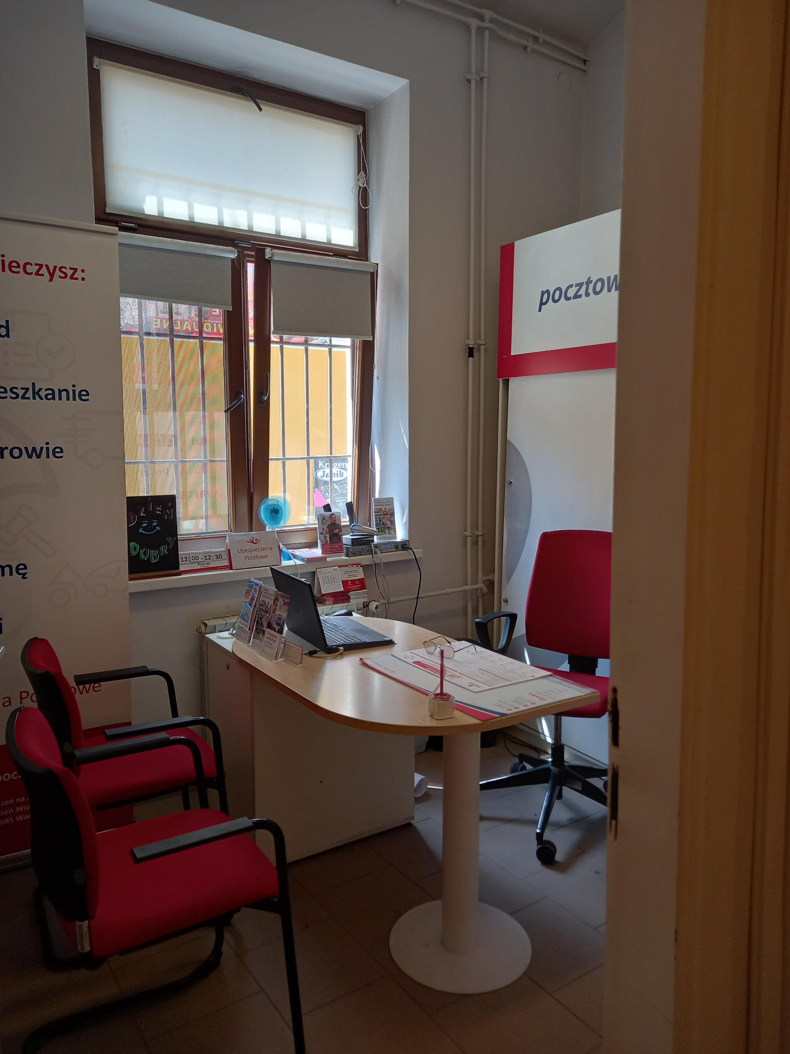 Biuro obsługi klienta z biurkiem, laptopem, krzesłami i plakatem z napisem 'ubezpieczenia pocztowe' przy oknie.