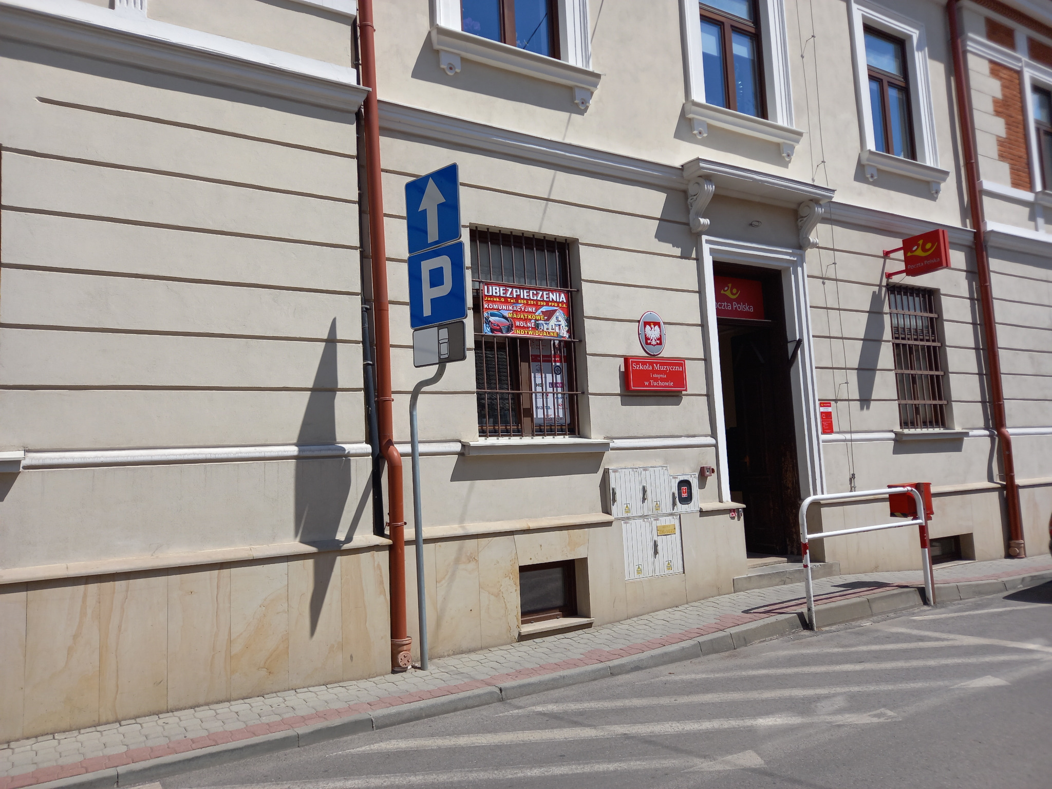 Budynek Poczty Polskiej z oknem, na którym widnieją plakaty reklamujące ubezpieczenia komunikacyjne, majątkowe, rolne i indywidualne. Obok okna znak drogowy 'parking'.