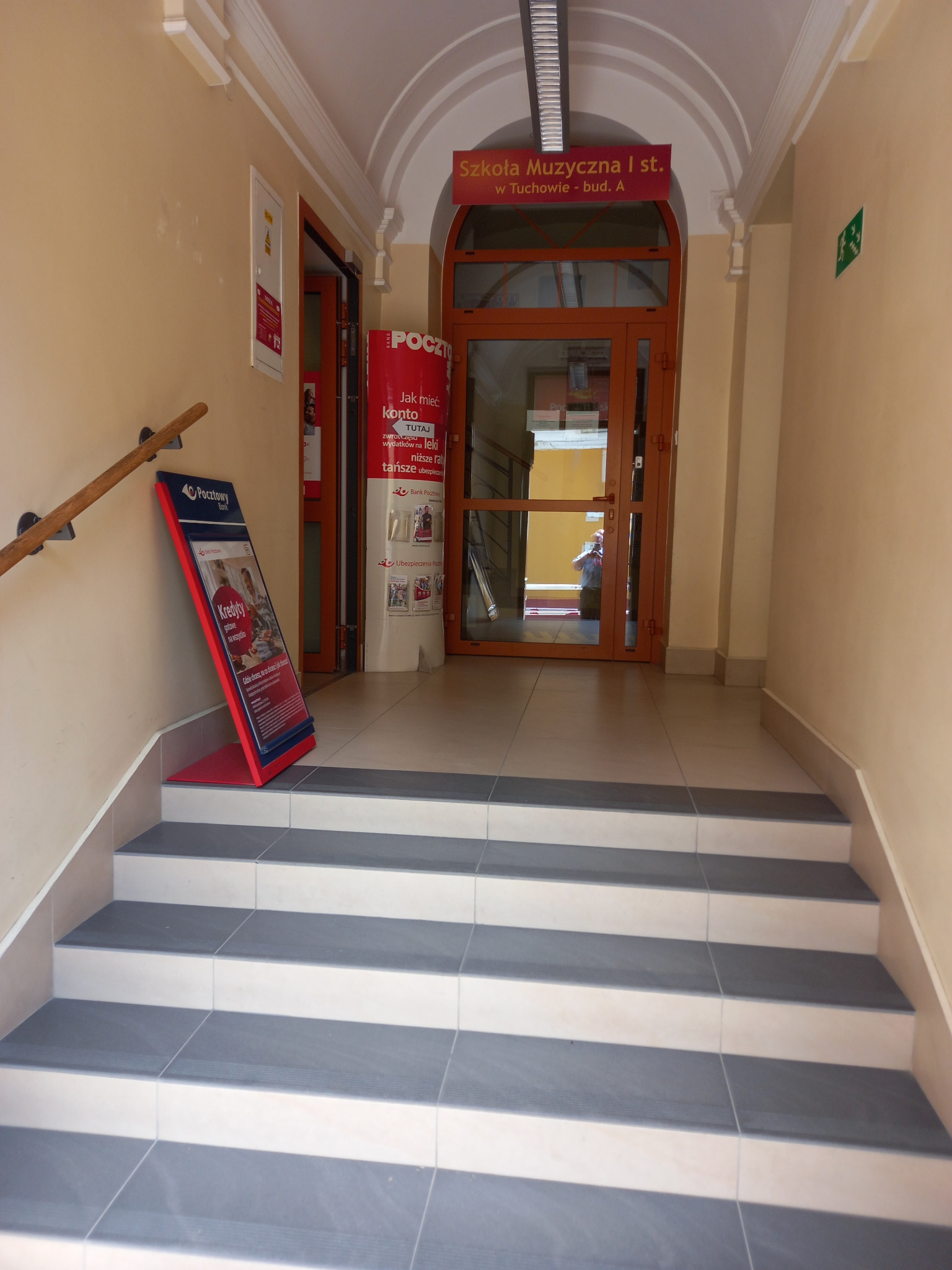 Wnętrze budynku z widocznymi schodami prowadzącymi do wejścia oznaczonego jako Szkoła Muzyczna I st. w Tuchowie. Po lewej stronie schodów stoi stojak reklamowy Banku Pocztowego z ofertą kredytów...