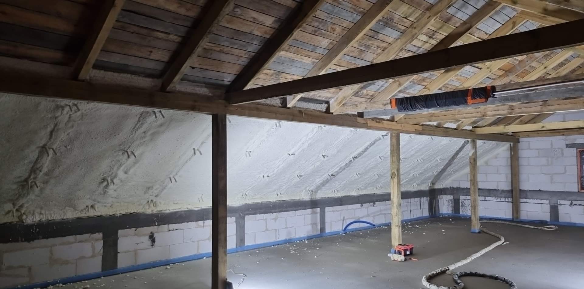 Poddasze ocieplone pianką PUR, widoczne drewniane krokwie, biała pianka na ścianach i skośnym dachu, oraz fragment podłogi z niebieską folią zabezpieczającą.