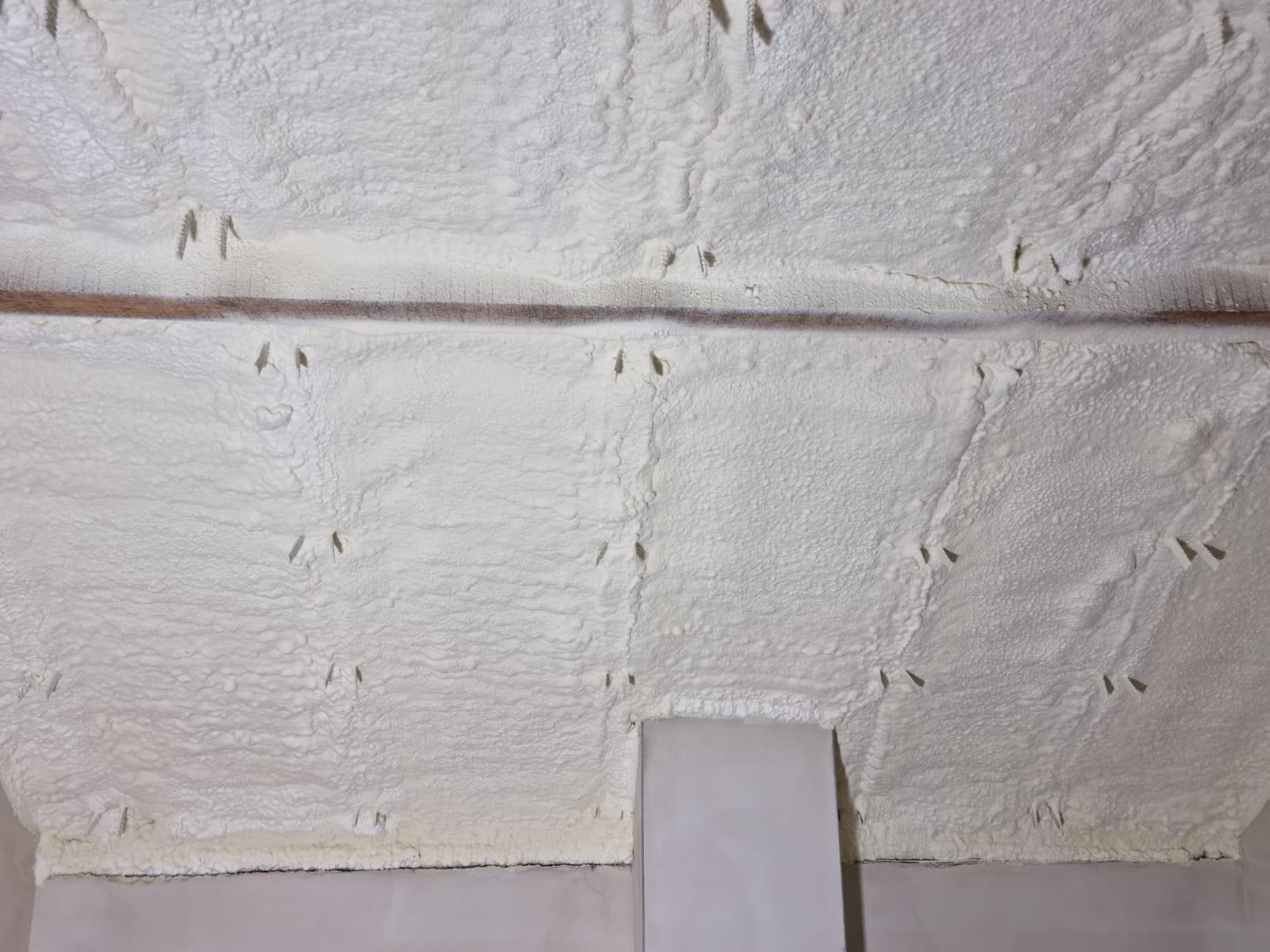 Poddasze ocieplone białą pianką poliuretanową, widoczne ślady po kołkach montażowych i drewniana belka stropowa.