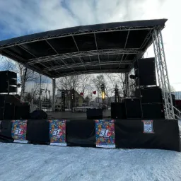 Zewnętrzna scena z nagłośnieniem i oświetleniem przygotowana na imprezę plenerową, widoczne plakaty z datą 31 grudnia, na śniegu.