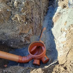 Pomarańczowe rury kanalizacyjne w wykopie, widoczne połączenie trójnikowe i fragment rury w ziemi, pionowe ściany wykopu z widocznymi korzeniami.
