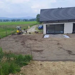 Żółta mini koparka przygotowuje teren pod budowę domu w Czernichowie. W tle widoczny pracownik z taczką, częściowo wykopana ziemia, a w oddali rozległe pola i góry.