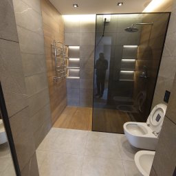 Apriori Władysław Arapov - Nowoczesna łazienka z szarymi płytkami imitującymi kamień, drewnopodobną ścianą z grzejnikiem drabinkowym i podświetlanymi niszami oraz kabiną prysznicową ze szkła.