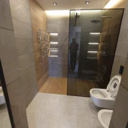 Nowoczesna łazienka z szarymi płytkami imitującymi kamień, drewnopodobną ścianą z grzejnikiem drabinkowym i podświetlanymi niszami oraz kabiną prysznicową ze szkła.