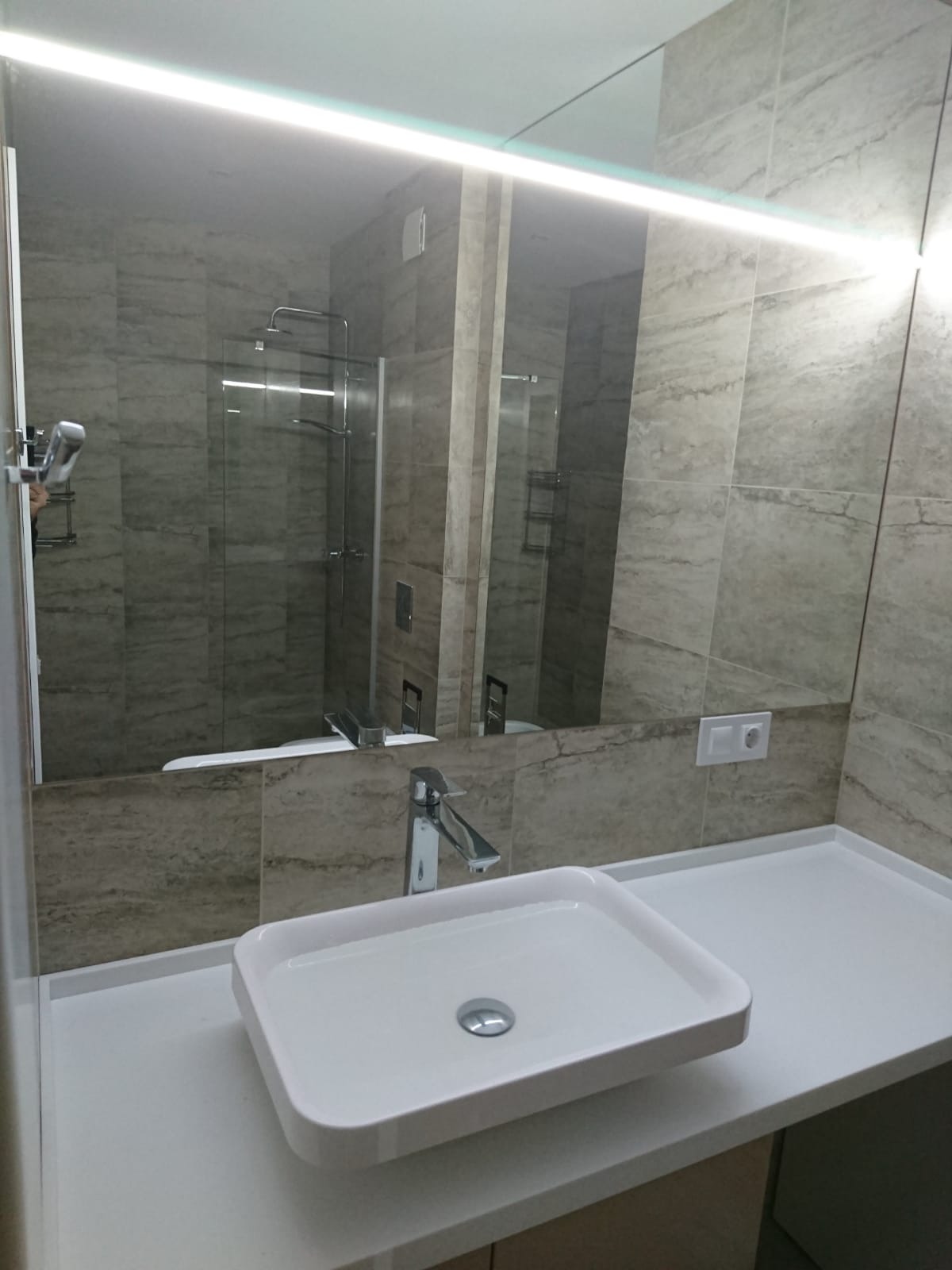 Nowoczesna łazienka z prostokątną umywalką nablatową, baterią chromowaną i dużym lustrem z podświetleniem LED, w tle widoczna kabina prysznicowa z szklanymi drzwiami.