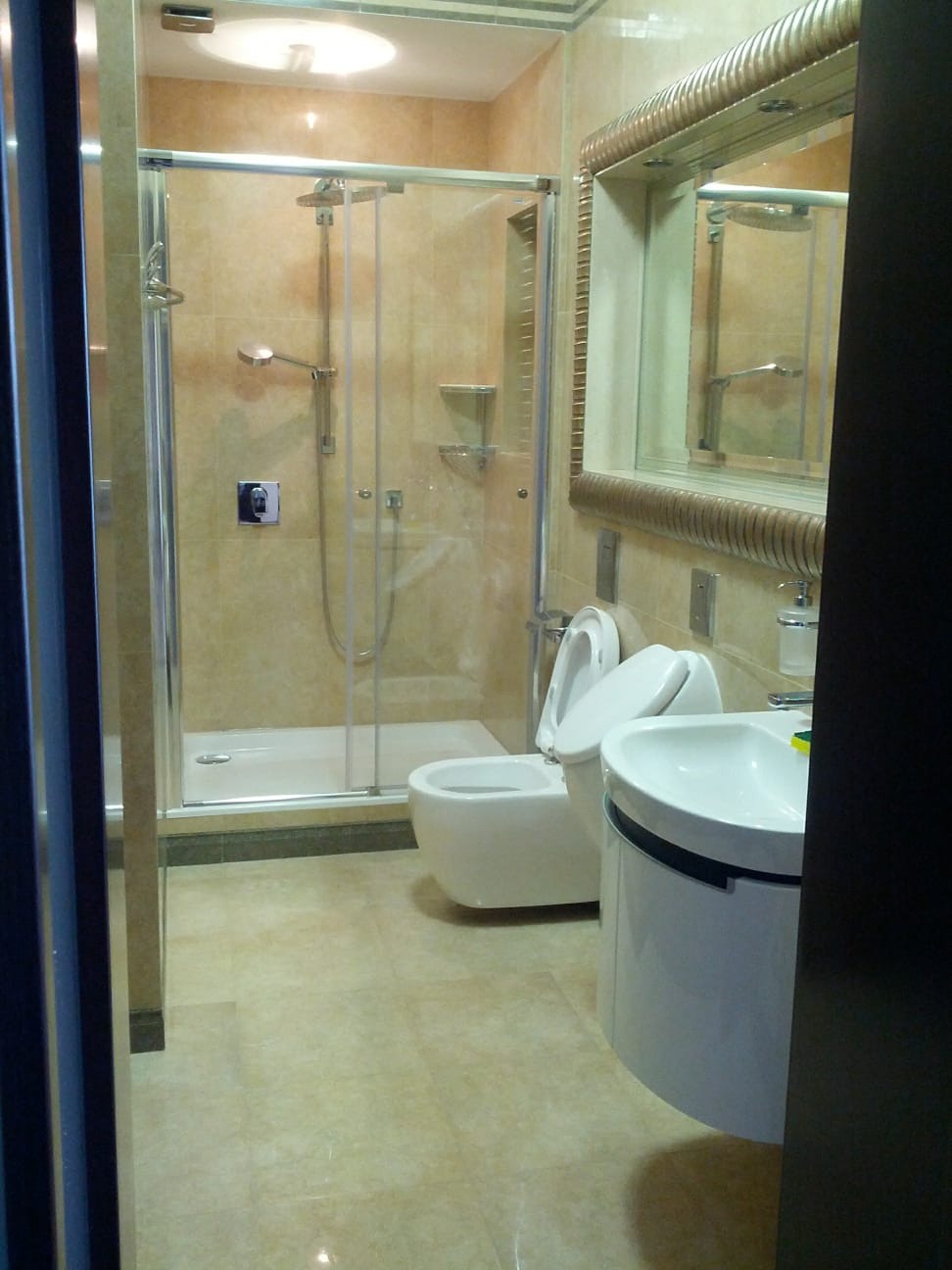 Wykończona łazienka z kabiną prysznicową, toaletą i umywalką, utrzymana w beżowej kolorystyce z lustrem w ozdobnej ramie.