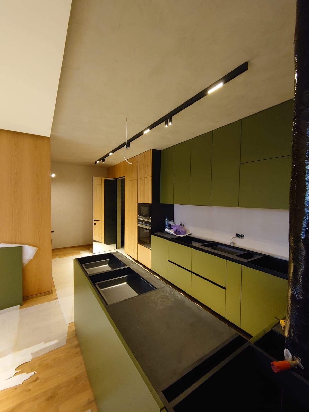 Wnętrze kuchni w trakcie montażu: zielone szafki kuchenne z czarnymi blatami, drewniane wykończenia, oświetlenie szynowe podsufitowe, podłoga zabezpieczona folią malarską.