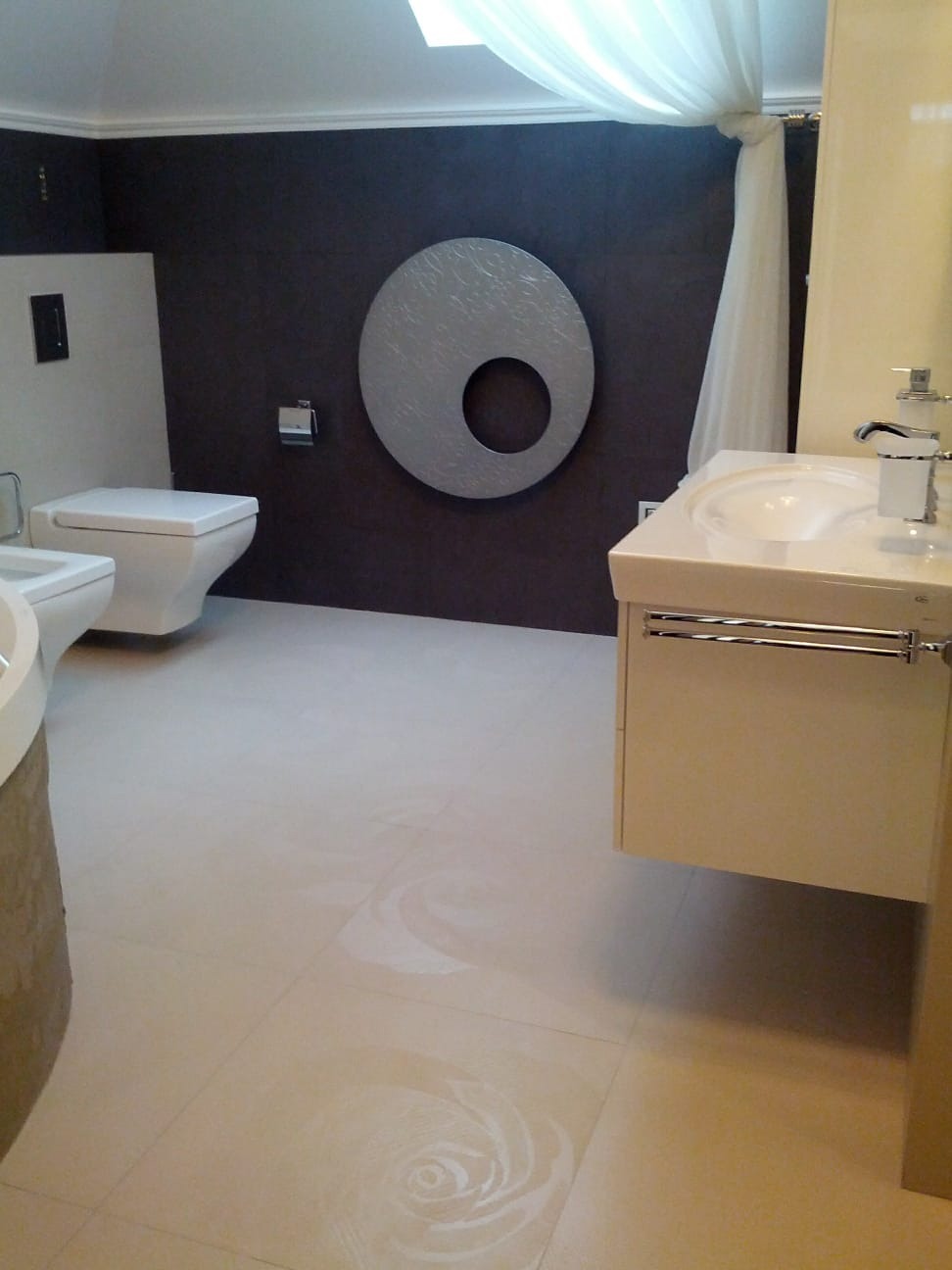 Wykończona łazienka z beżowymi płytkami podłogowymi z motywem róży, ciemnobrązową ścianą z okrągłą dekoracją i białymi zasłonami pod sufitem.