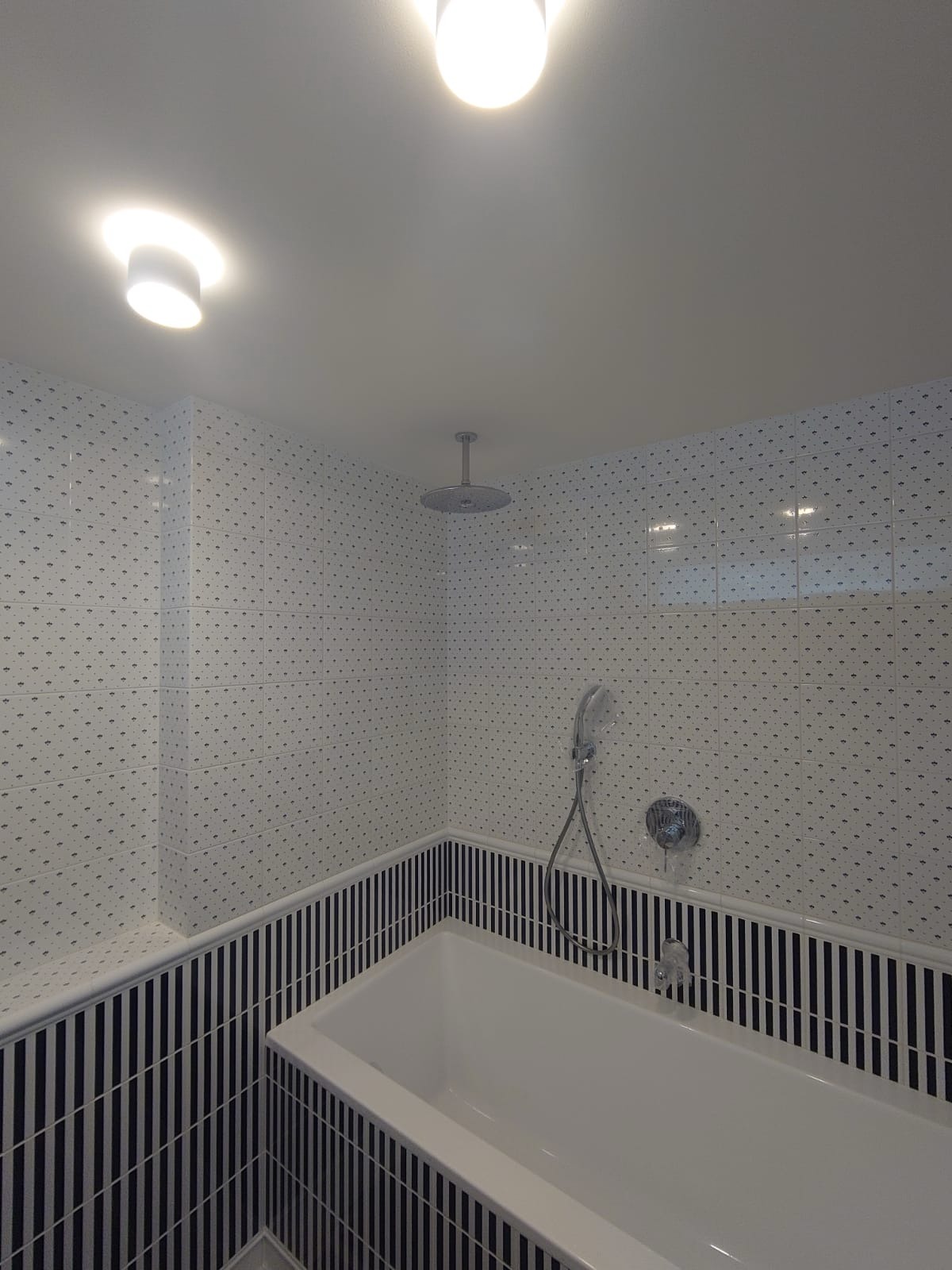 Biała wanna w narożniku łazienki wyłożonej płytkami w czarno-białe paski i kropki, widoczna bateria i deszczownica.