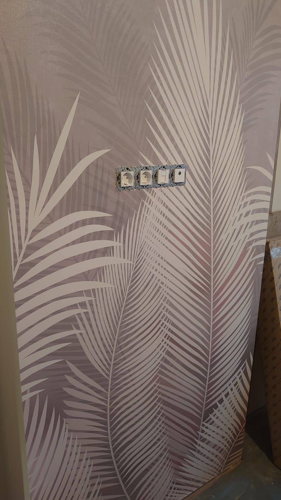 Ściana z tapetą w motywie liści palmowych w odcieniach szarości, z widocznymi gniazdkami elektrycznymi i włącznikami bez obudowy, w trakcie prac wykończeniowych.