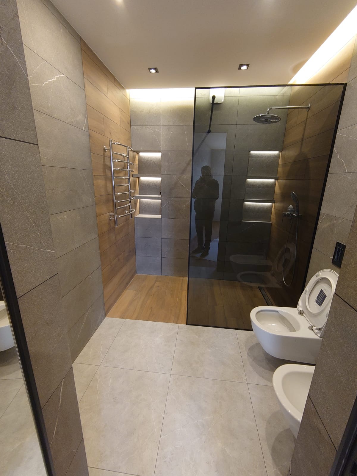 Nowoczesna łazienka z szarymi płytkami imitującymi kamień, drewnopodobną ścianą z grzejnikiem drabinkowym i podświetlanymi niszami oraz kabiną prysznicową ze szkła.