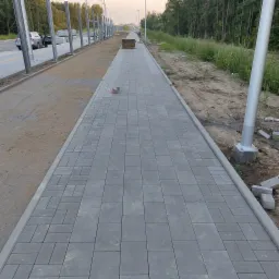 Nowy chodnik z szarej kostki brukowej ułożonej we wzór, obrzeża z krawężników, w tle słupy oświetleniowe i drzewa.