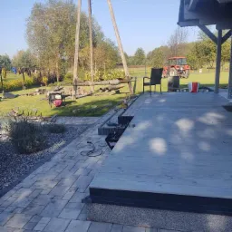 Wiercenie studni na trawniku z widocznymi rurami wiertniczymi, agregatem prądotwórczym oraz traktorem w tle, ujęcie z poziomu tarasu z kostki brukowej.