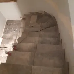 Schody wyłożone płytkami w trakcie montażu, widoczne krzyżyki dystansowe i puszki z pianką montażową, fragment podłogi z ułożonymi płytkami.