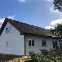 Nowoczesny dom jednorodzinny z białymi ścianami i ciemnobrązowym dachem, widok z zewnątrz w słoneczny dzień z niebieskim niebem i chmurami, w tle drzewa.