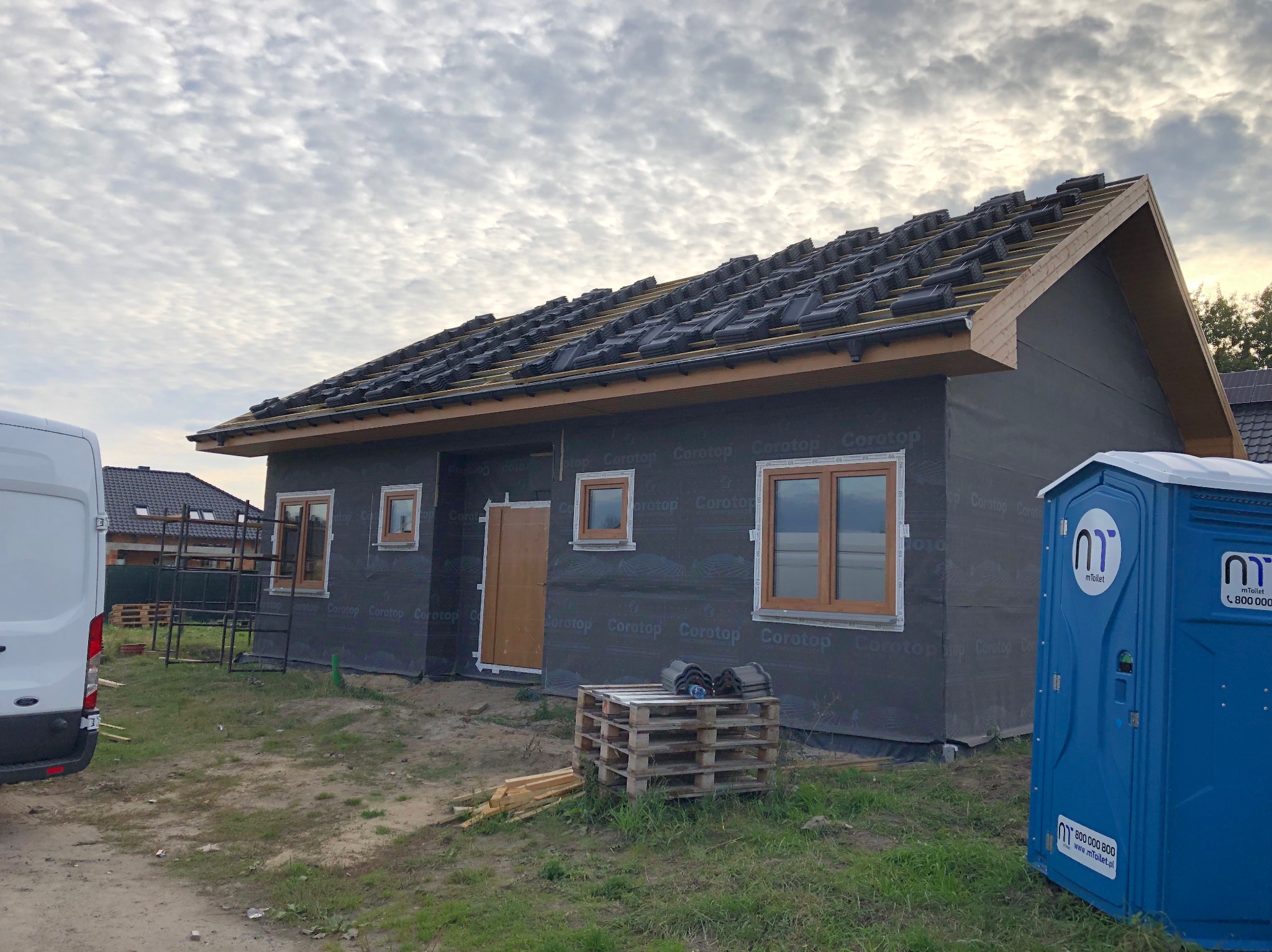 Budowa domu w Golczewie na etapie kładzenia dachu, widoczna membrana na ścianach i okna w drewnianych ramach. Na pierwszym planie przenośna toaleta i paleta z dachówkami.