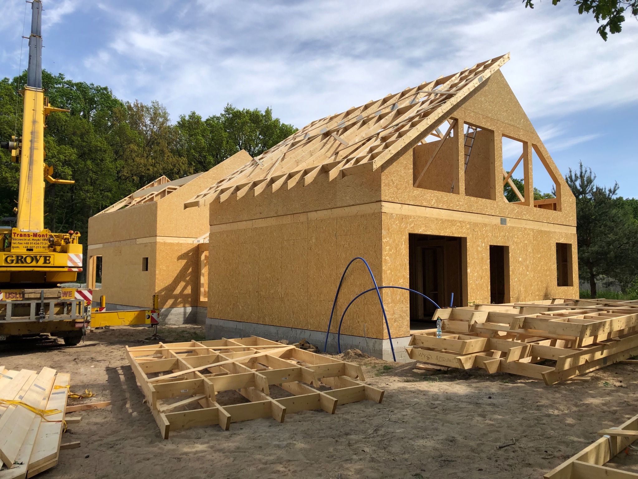 Budowa domu w technologii szkieletowej w trakcie montażu konstrukcji dachu, z widocznym żurawiem budowlanym i przygotowanymi elementami podłogi.