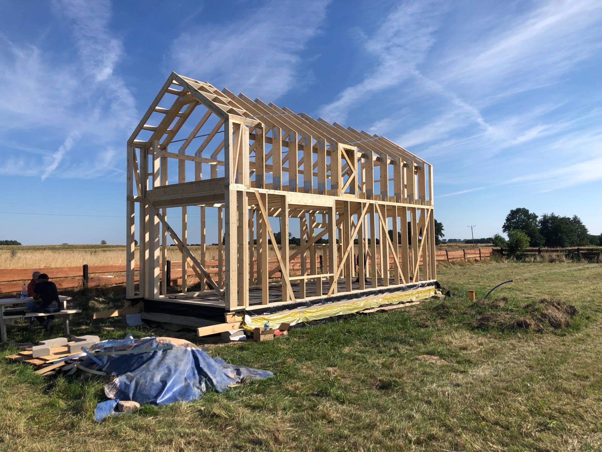 Konstrukcja szkieletowa domu w trakcie budowy, widoczne drewniane elementy ramy na fundamencie z hydroizolacją, w tle łąka i błękitne niebo z chmurami, obok plac budowy z materiałami i pracownikami.