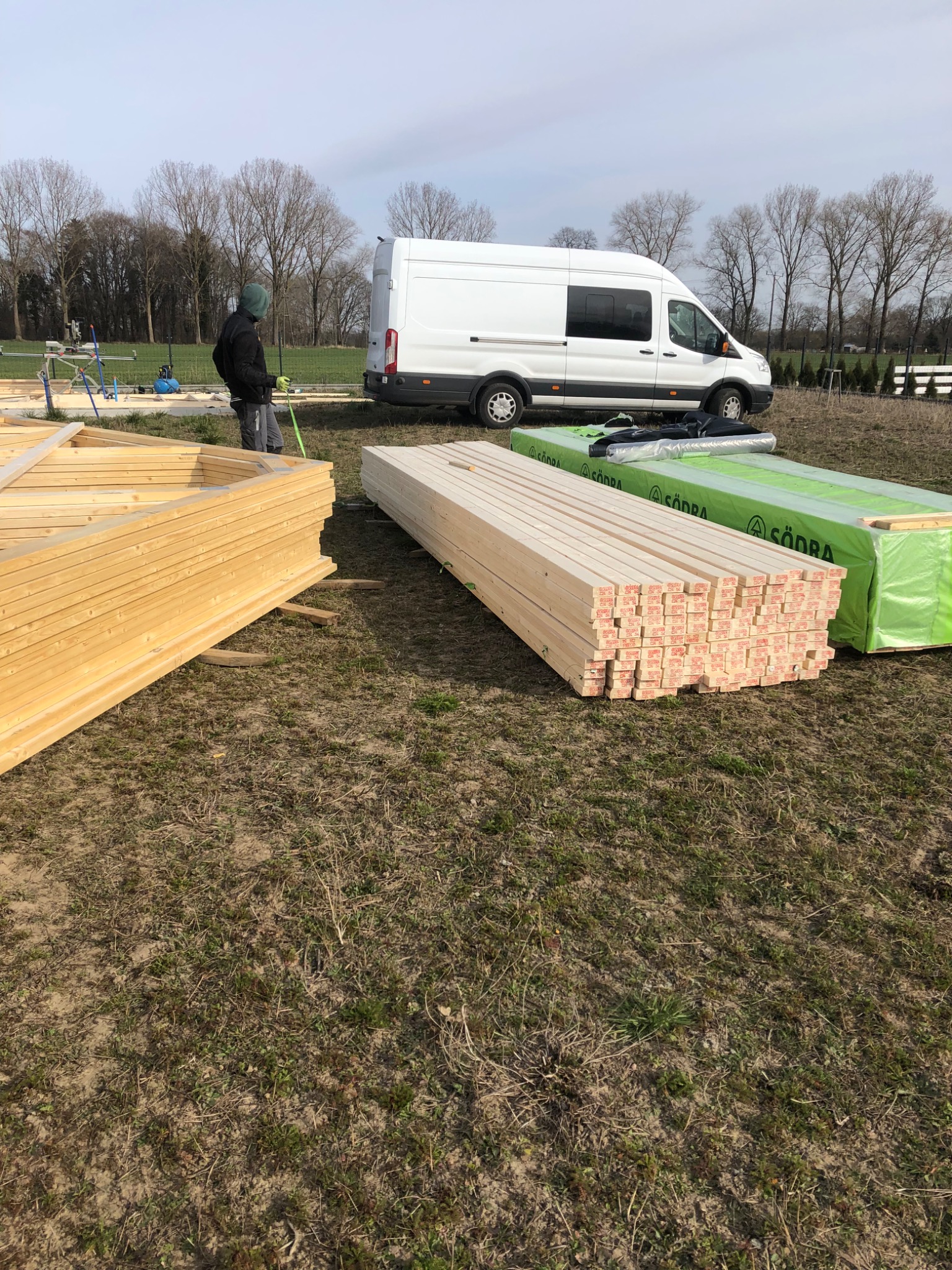 Przygotowane elementy konstrukcyjne z drewna i zapakowane materiały izolacyjne na placu budowy, biały samochód dostawczy w tle, pracownik mierzy element konstrukcji.