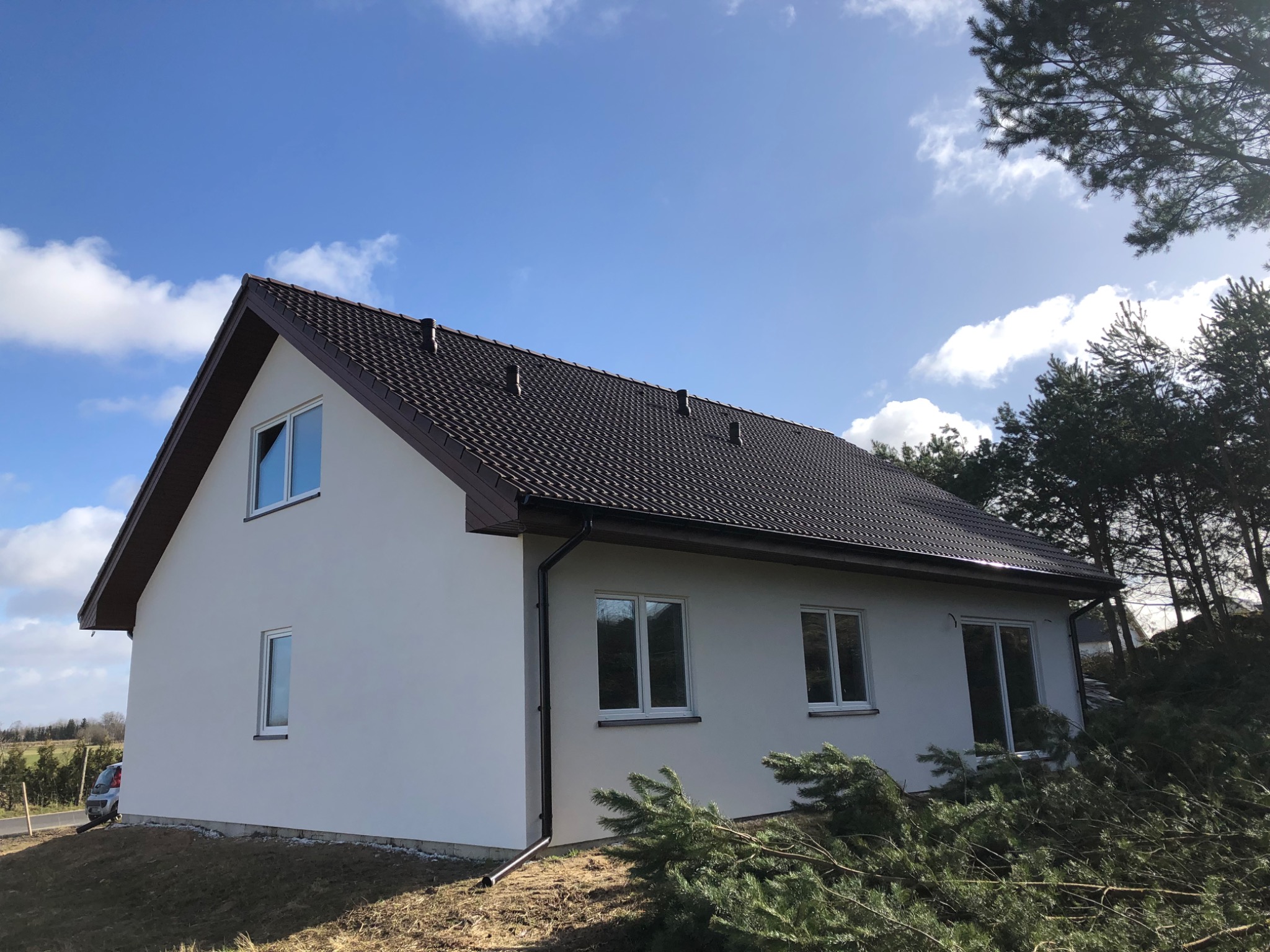 Nowoczesny dom jednorodzinny z białymi ścianami i ciemnobrązowym dachem, widok z zewnątrz w słoneczny dzień z niebieskim niebem i chmurami, w tle drzewa.