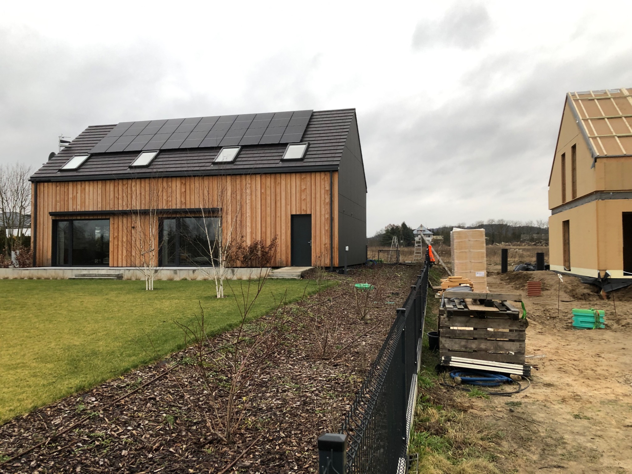 Nowoczesny dom z drewnianą elewacją i panelami słonecznymi na dachu, obok budynek w trakcie budowy, widoczne materiały budowlane i ogrodzenie.