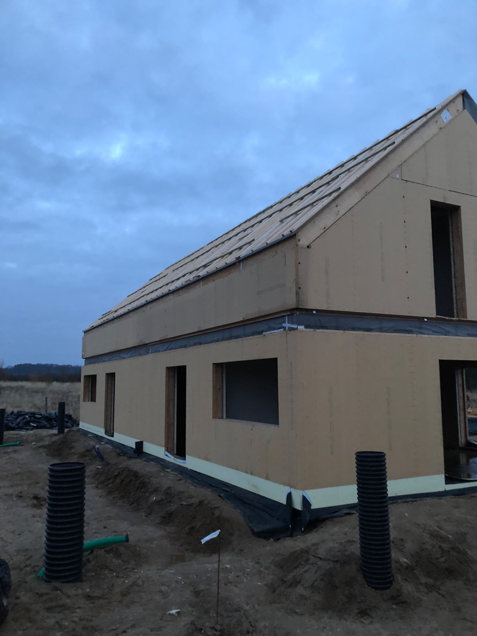 Surowy stan dwukondygnacyjnego domu szkieletowego w trakcie budowy, widoczne panele ścienne, przygotowana konstrukcja dachu i czarne rury drenażowe w ziemi.