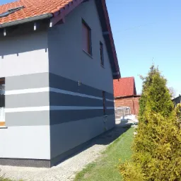 Szary dom jednorodzinny z pomalowaną elewacją w poziome pasy różnych odcieni szarości i bieli, widok z boku na tle zielonego trawnika i błękitnego nieba.