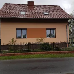 Dwukolorowa elewacja domu jednorodzinnego z brązową wstawką, widok z poziomu ulicy, czarne ogrodzenie z ceglaną podmurówką, czerwona dachówka i dwa okna dachowe.