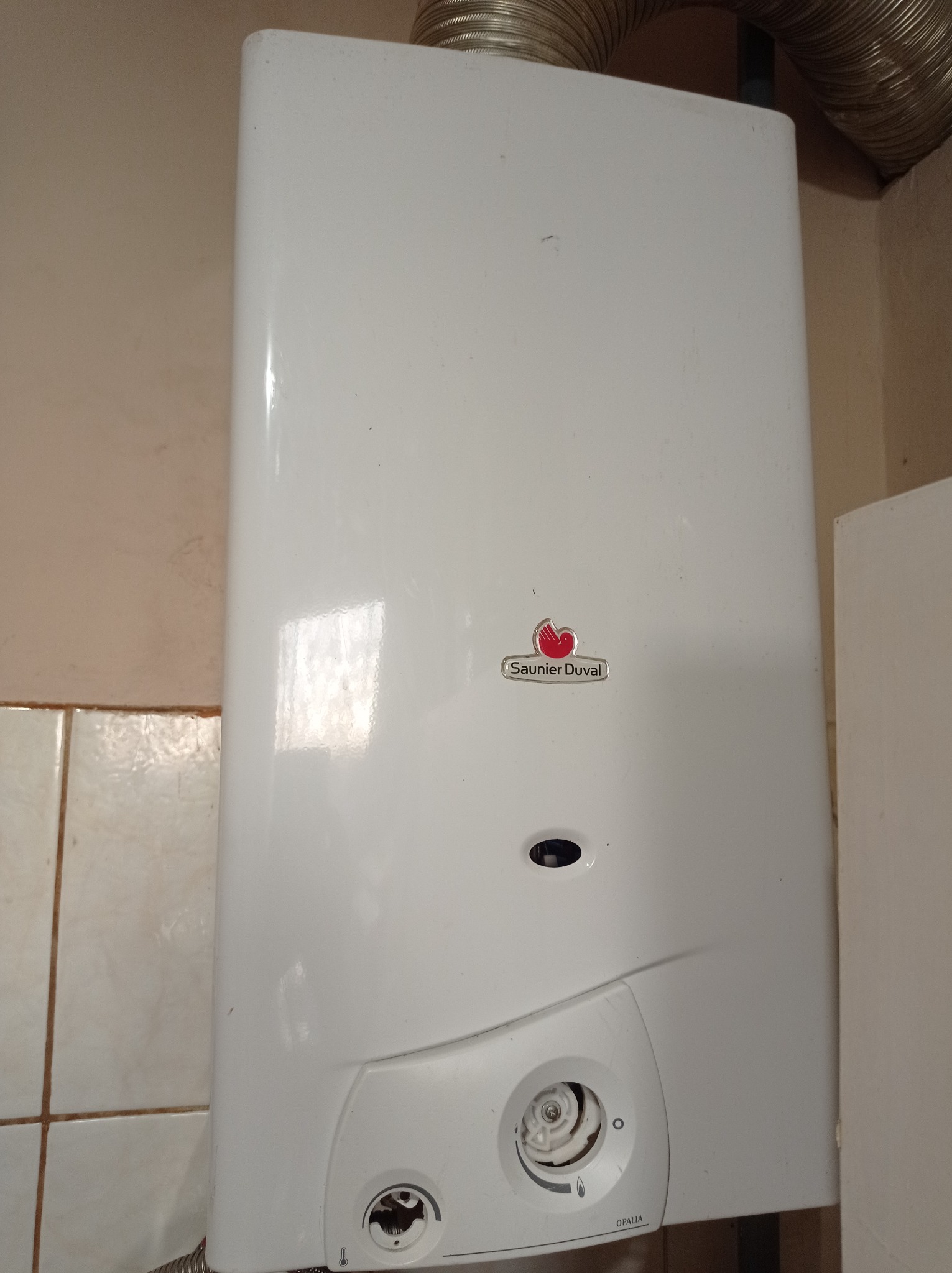 Biały, wiszący piec gazowy Saunier Duval Opalia z widoczną rurą odprowadzającą spaliny i fragmentem ściany z beżowymi płytkami w tle.