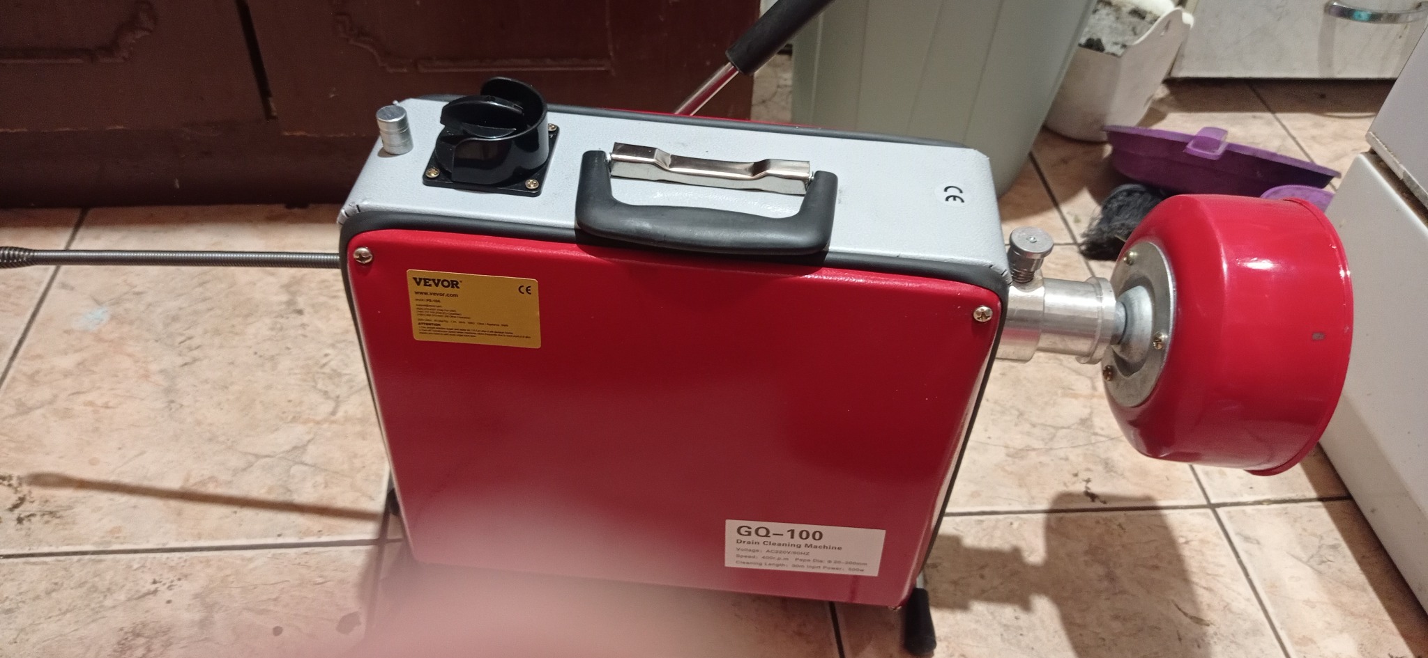 Przenośna elektryczna maszyna do czyszczenia rur VEVOR GQ-100 z elastycznym wężem spiralnym, stojąca na kafelkach.