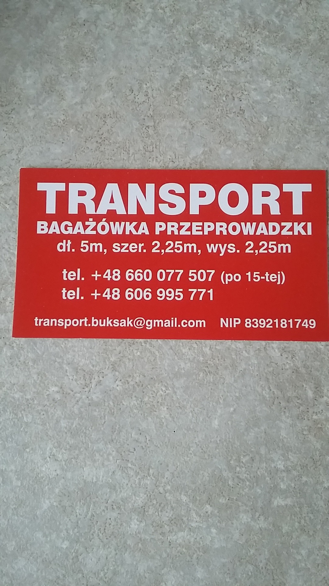 Czerwona wizytówka firmy transportowej z napisem 'TRANSPORT BAGAŻÓWKA PRZEPROWADZKI', wymiarami bagażówki, numerami telefonów i adresem e-mail, leżąca na szarej powierzchni.