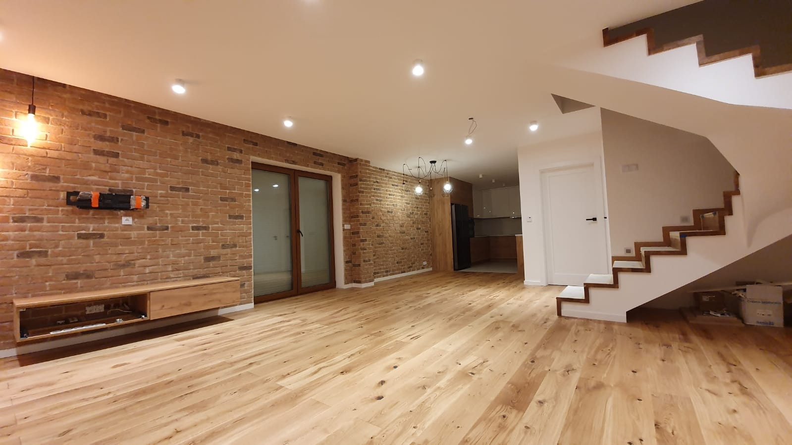 Przestronny salon z ceglaną ścianą, drewnianą podłogą i schodami z drewnianymi stopniami. Na ścianie widoczny uchwyt na telewizor. W głębi widoczna kuchnia.