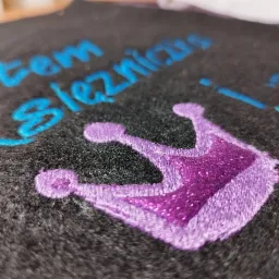 Czarny materiał z wyhaftowanym fioletowym brokatowym wzorem korony i niebieskim napisem 'Jestem Ksieżniczką', zbliżenie, widoczne detale ściegów.