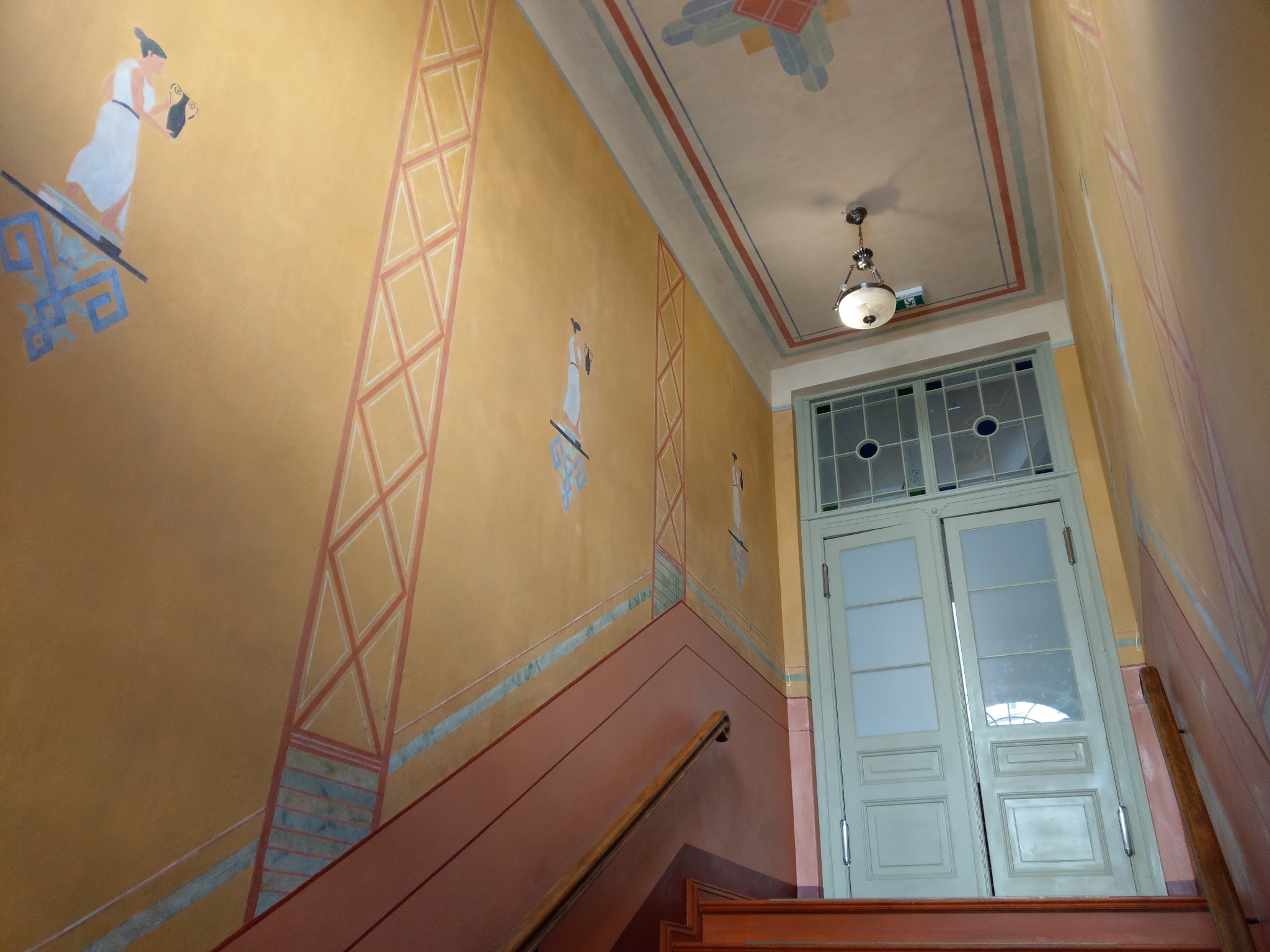 Art Deco klatka schodowa z malowidłami na ścianach przedstawiającymi postacie w stylu antycznym, geometryczne wzory i dwuskrzydłowe drzwi z przeszkleniami.
