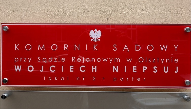 Czerwona tabliczka informacyjna z napisem 'Komornik Sądowy przy Sądzie Rejonowym w Olsztynie Wojciech Niepsuj lokal nr 2 parter', umieszczona na ścianie.
