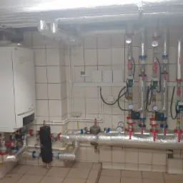 Kotłownia z białym piecem gazowym, rurami pokrytymi folią izolacyjną, manometrami i zaworami na kafelkowej ścianie i podłodze.