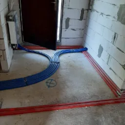 Wnętrze w trakcie prac instalacyjnych: nieotynkowane ściany z betonu komórkowego, podłoga w surowym betonie, ułożone równolegle niebieskie rury instalacji wodnej i czerwone rury grzewcze wzdłuż...