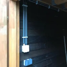 Szare rury instalacji elektrycznej przymocowane do drewnianego stropu i czarnej, drewnianej ściany, z widoczną puszką elektryczną i podwójnym włącznikiem.