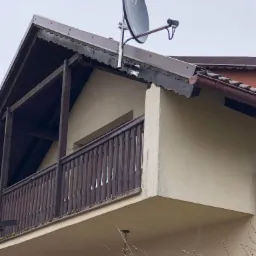 Antena satelitarna zamontowana na dachu domu z drewnianym balkonem, widok z dołu na tle pochmurnego nieba.