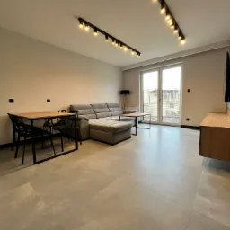 Nowoczesny salon z szarą sofą narożną, drewnianym stołem i czarnymi krzesłami, oświetleniem punktowym na suficie i dużymi oknami balkonowymi.