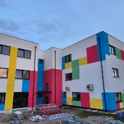 Nowoczesny budynek po malowaniu elewacji w geometryczne, kolorowe wzory. Widoczne rusztowania i teren po budowie. Oświetlenie wieczorne.