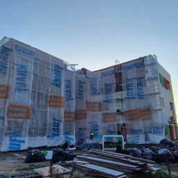 REMONTY & WNĘTRZA Piotr Piestrzyński - Budynek w trakcie ocieplania, osłonięty siatką z logo Knauf, rusztowania, widoczne materiały budowlane. Prace w toku, fragment elewacji z kolorem.