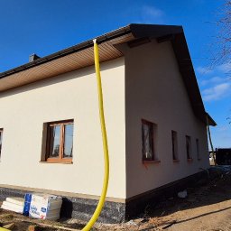 REMONTY & WNĘTRZA Piotr Piestrzyński - Jasny dom jednorodzinny po tynkowaniu, z widocznymi oknami zabezpieczonymi folią. Na pierwszym planie żółty wąż i styropian. Błękitne niebo w tle.