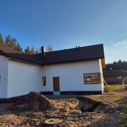 REMONTY & WNĘTRZA Piotr Piestrzyński - Nowoczesny dom jednorodzinny z białą elewacją, ciemnym dachem i brązowymi oknami, w trakcie prac wykończeniowych, z wykopem i stertą ziemi na pierwszym planie.