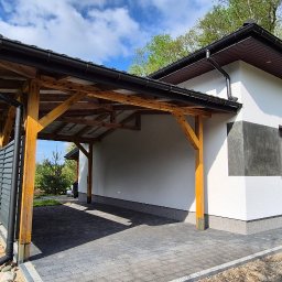 REMONTY & WNĘTRZA Piotr Piestrzyński - Nowoczesna elewacja domu z drewnianą konstrukcją wiaty garażowej i dekoracyjnym tynkiem w odcieniach szarości. Widok z zewnątrz, w tle zieleń drzew.