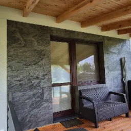 Wykończony balkon z drewnianym sufitem i ścianą z dekoracyjną szarą okładziną imitującą kamień. Widoczne meble ogrodowe i zieleń za barierką.