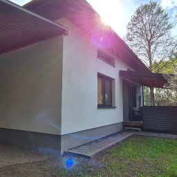 REMONTY & WNĘTRZA Piotr Piestrzyński - Jasny budynek z nową elewacją i podbitką dachową, widok z boku z fragmentem terenu budowy.  Na pierwszym planie trawnik. W tle drzewa i niebo.